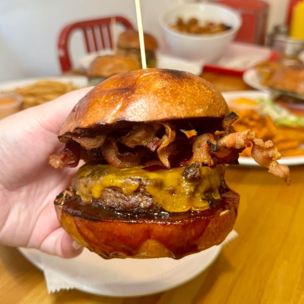 A hand holds a P.S. Burgers bacon cheeseburger with melted cheese and BBQ sauce.