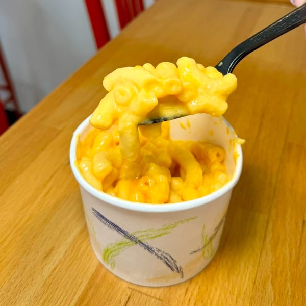 A black fork lifts creamy mac and cheese from a white cup on a wooden table.