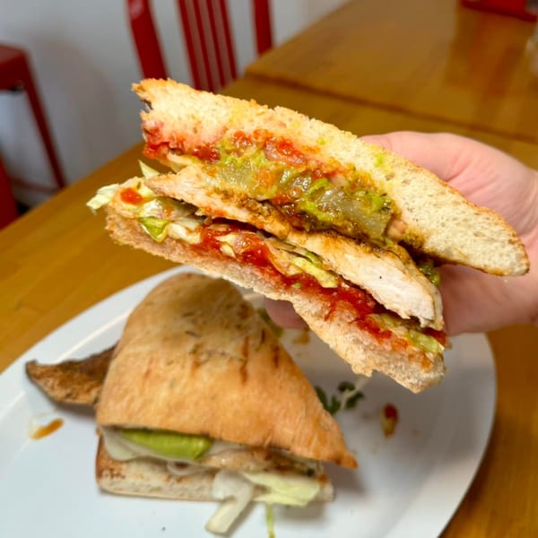 A hand holds up half of a chicken sandwich with red sauce and guacamole, another half rests on a plate.